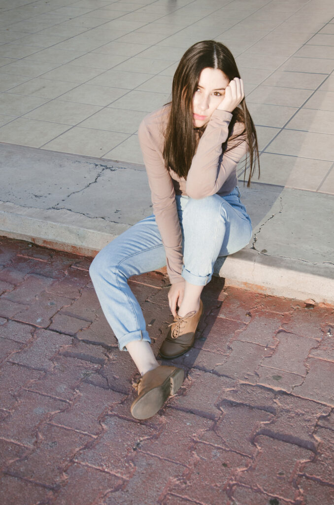 Gabriela Inzunza sits on a curb leaning on her hand wearing a brown top and blue jeans with brown shoes on.